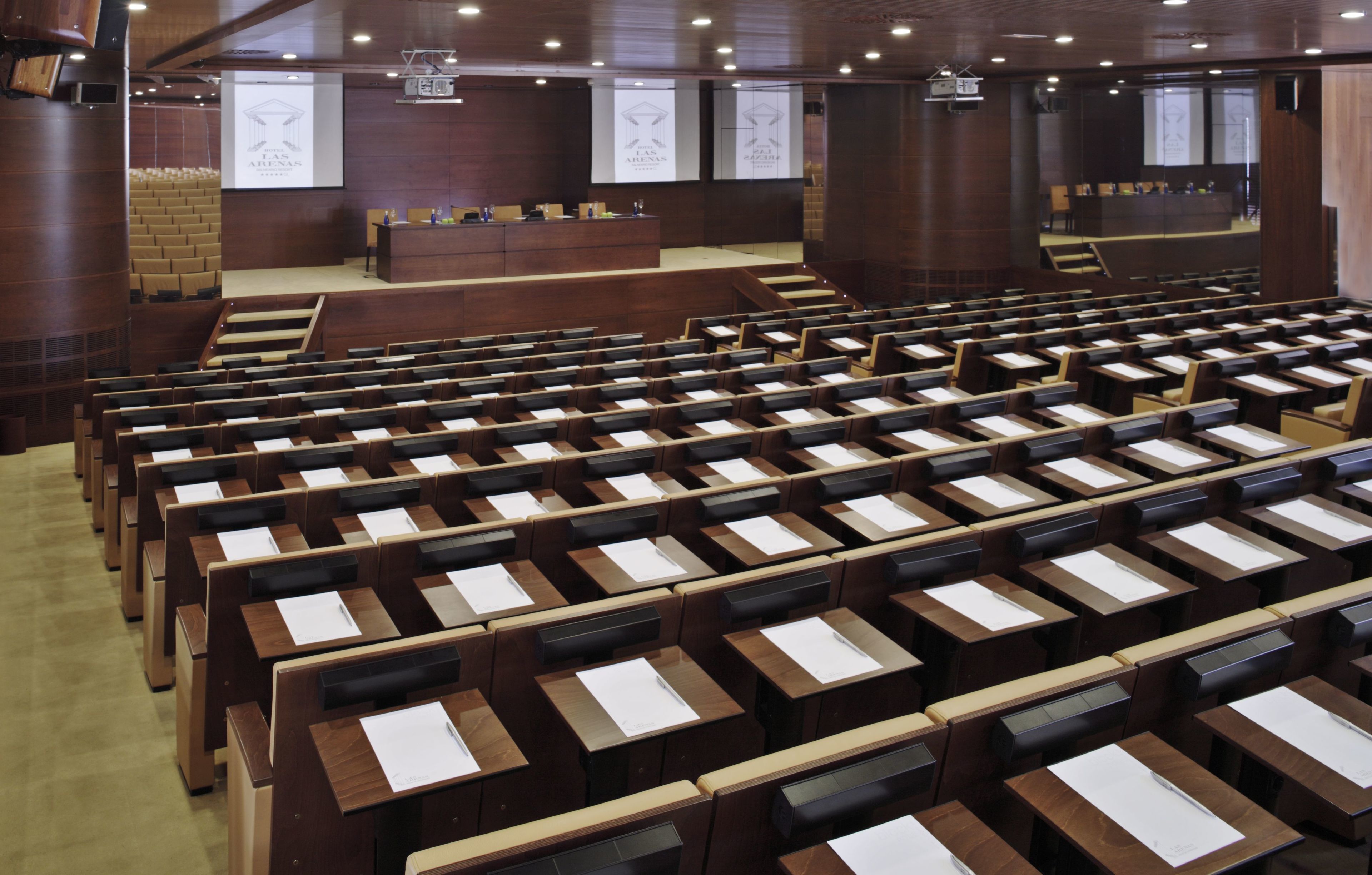 Un'ampia aula con file di banchi e sedie in legno, un leggio di fronte e uno schermo per proiettore.