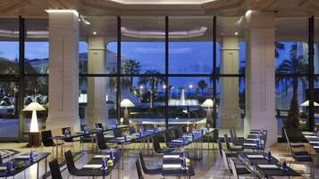 Un comedor amplio con mesas y sillas, ventanas grandes con vistas a palmeras y una fuente, e iluminación moderna.