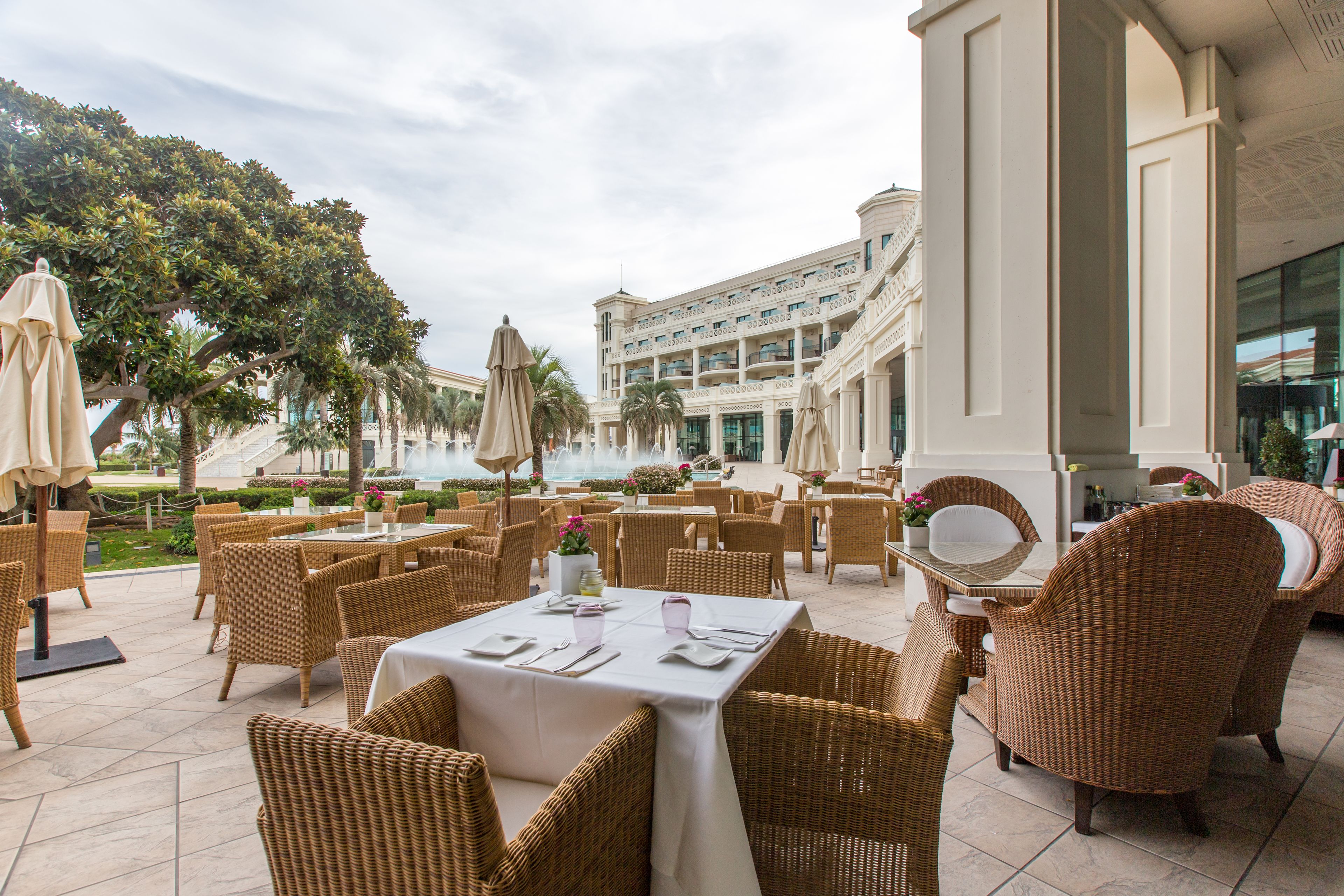 Un área de comedor al aire libre con mobiliario de mimbre y vistas a un edificio grande con varios pisos y balcones.