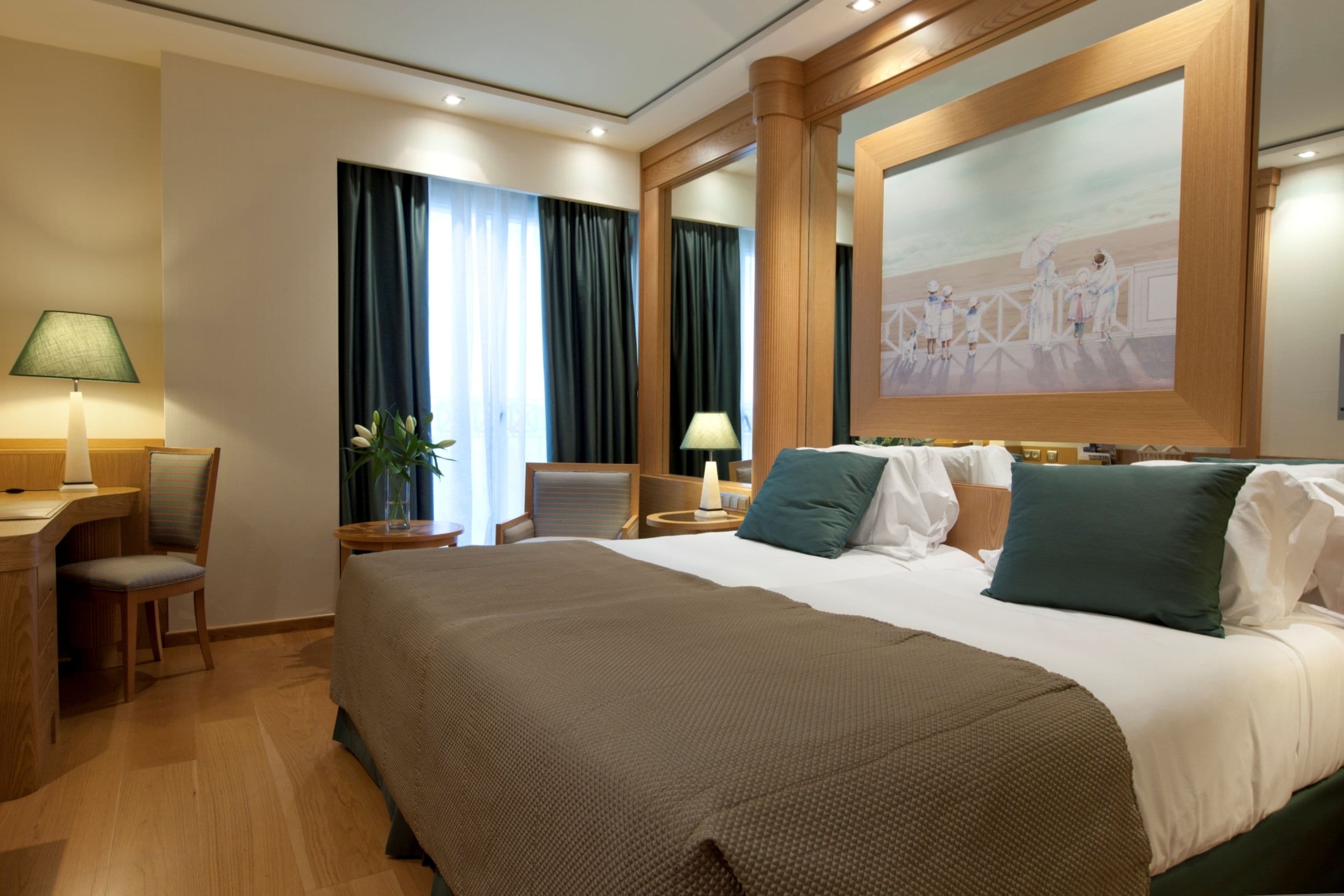 Ein Hotelzimmer mit einem großen Bett, einem Schreibtisch mit Lampe, einem Sessel, einem Nachttisch ebenfalls mit Lampe und einem Gemälde an der Wand.