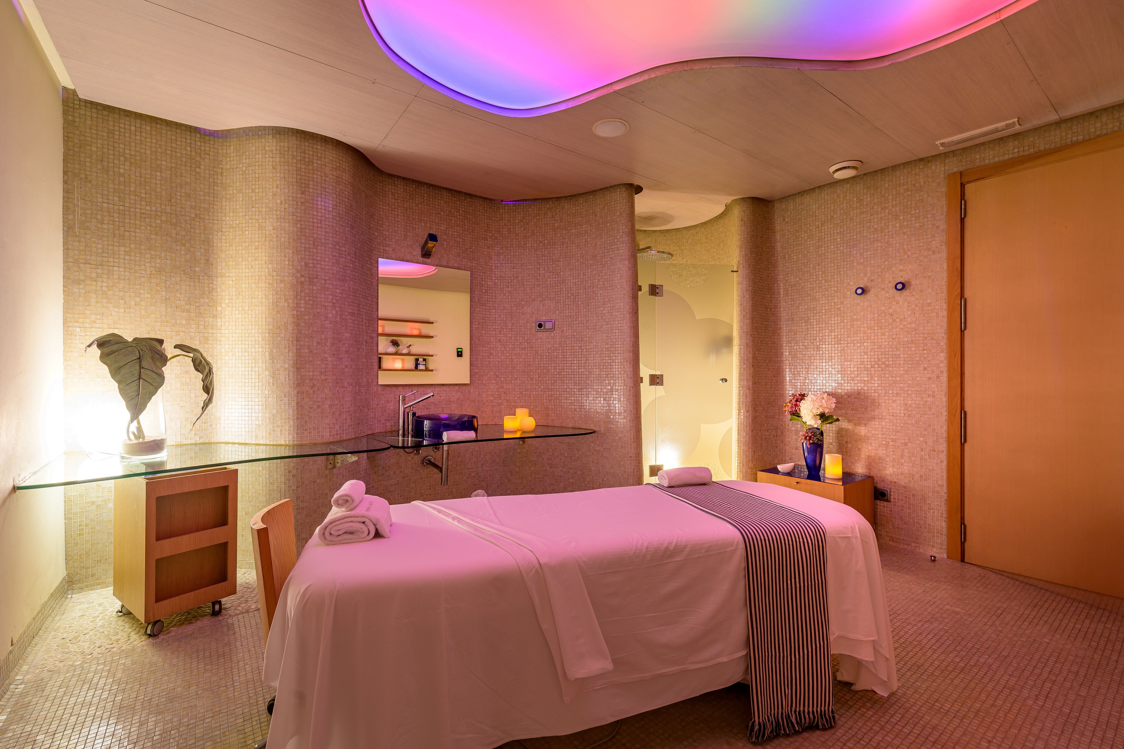 Habitación de hotel con cama, tocador con paredes de vidrio, escritorio pequeño y baño visible a través de una puerta de cristal.