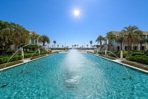 A large outdoor swimming pool with clear blue water, surrounded by palm trees and buildings.