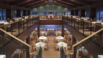 Un comedor amplio con varias mesas y sillas, una escalera central y ventanas grandes que ofrecen vistas al exterior.