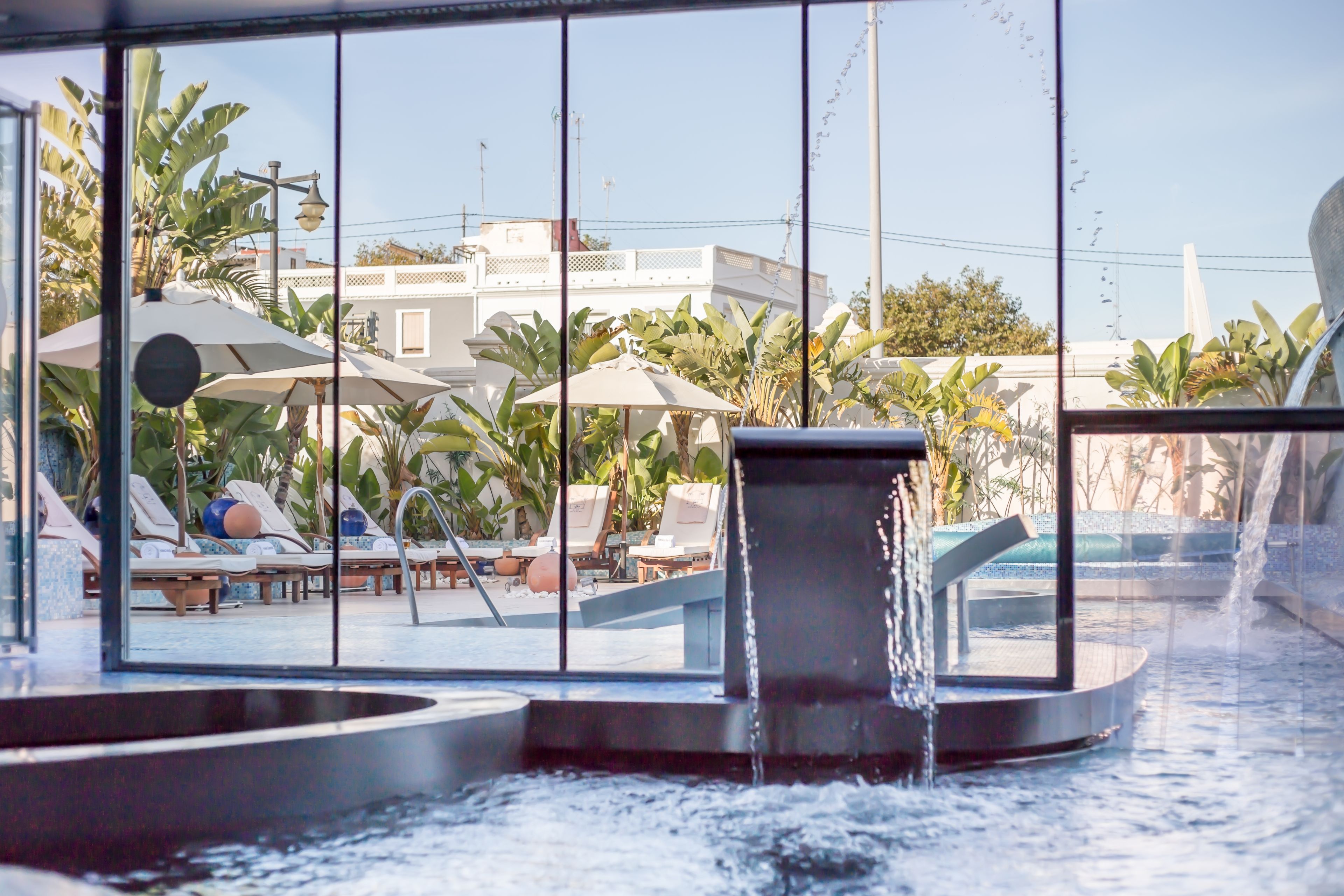 A pool area with lounge chairs and umbrellas, a water feature, and a view of buildings in the background.