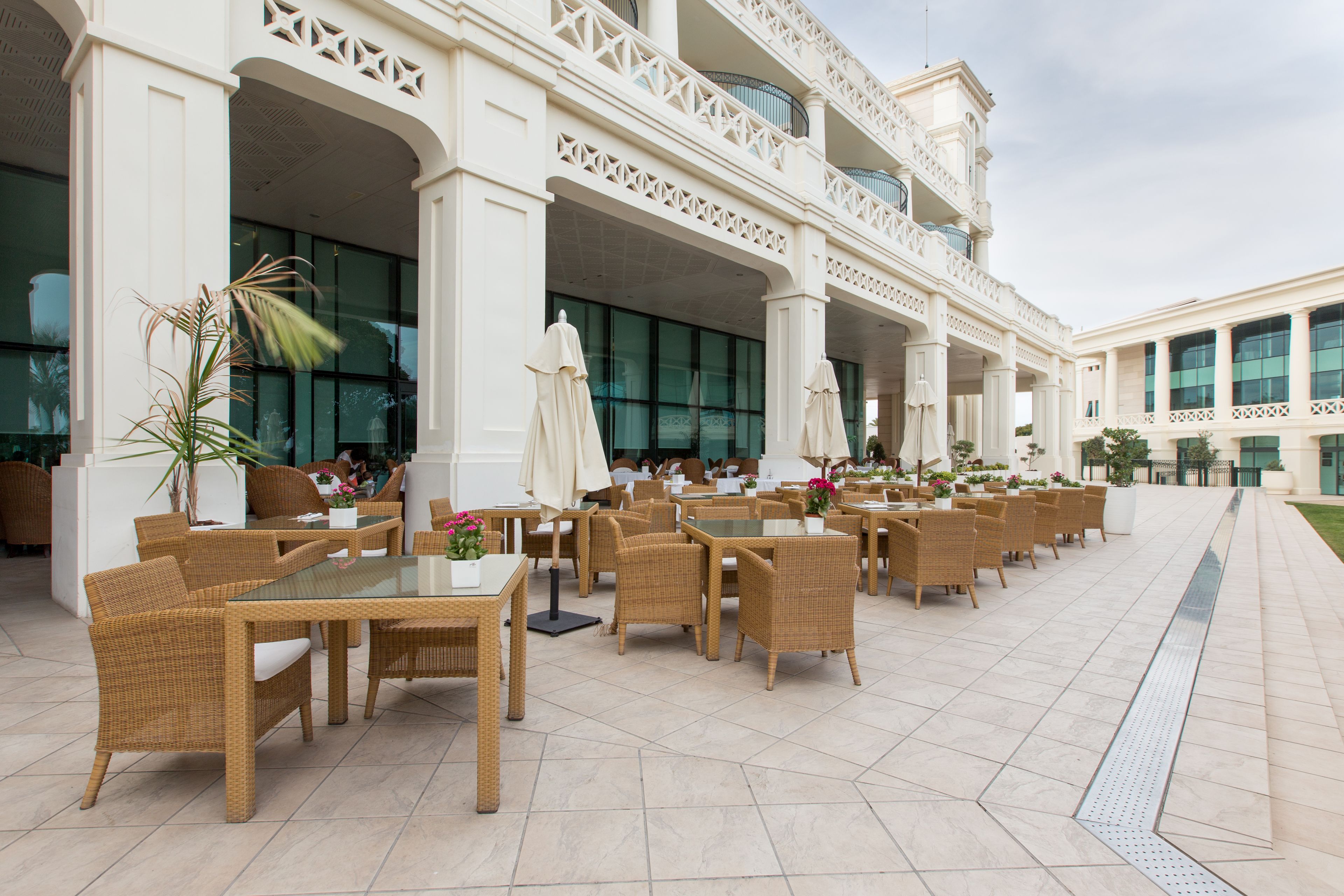 An outdoor seating area with wicker furniture and umbrellas, a tiled floor, and a building with large windows and decorative balconies.