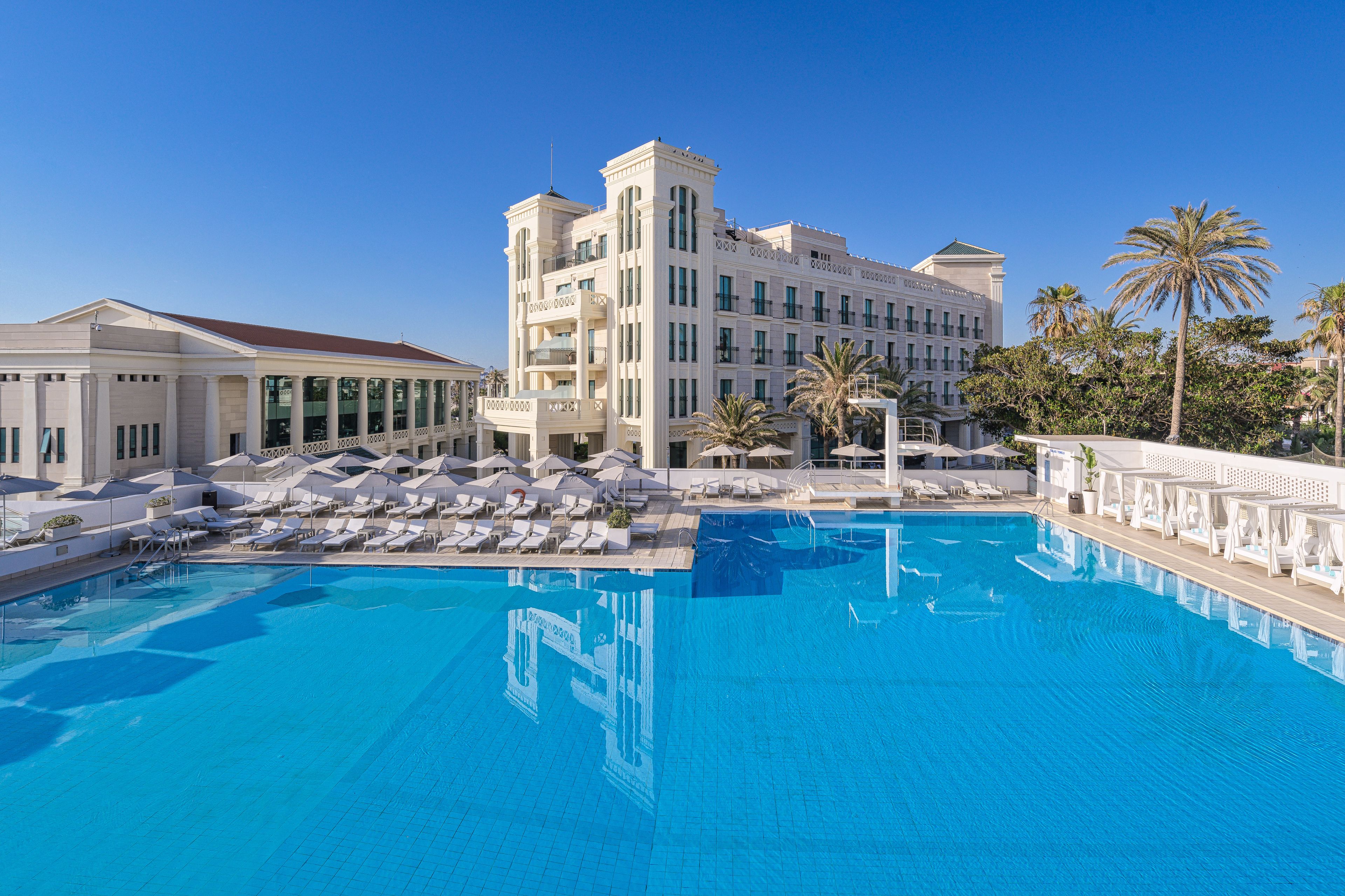Una amplia piscina con sillones de descanso y sombrillas, rodeada por un edificio de hotel de varios pisos con un estilo arquitectónico clásico.