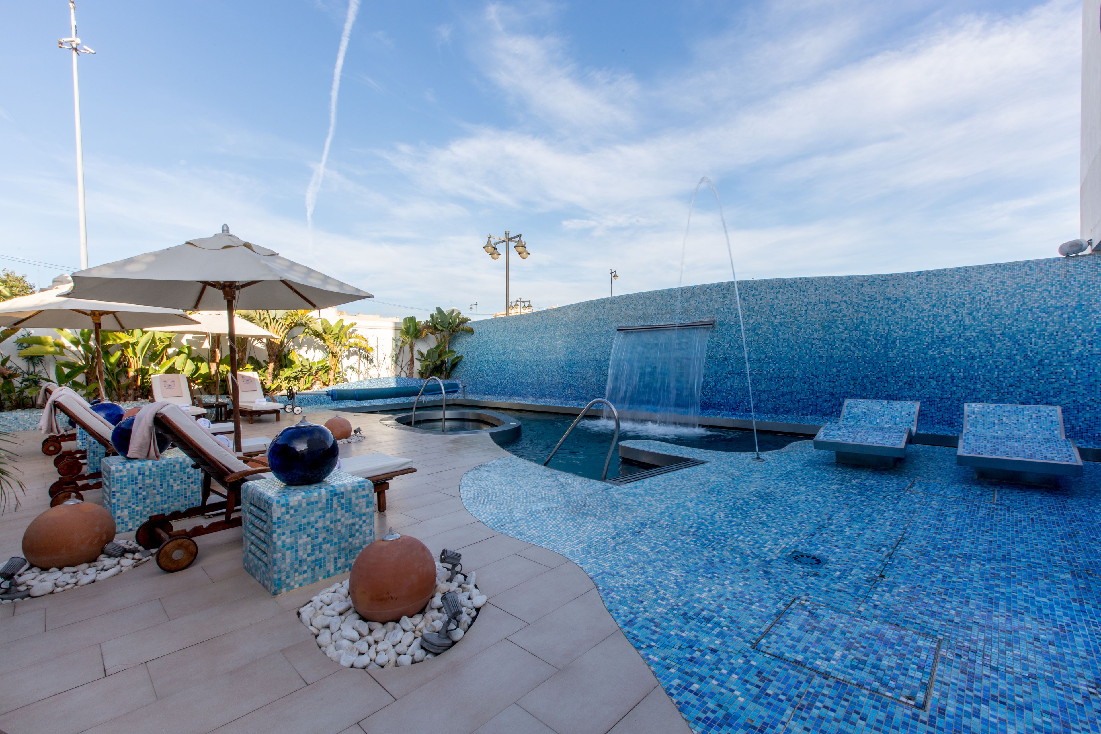 Zona de piscina con sillones de descanso, sombrillas y una fuente de agua.