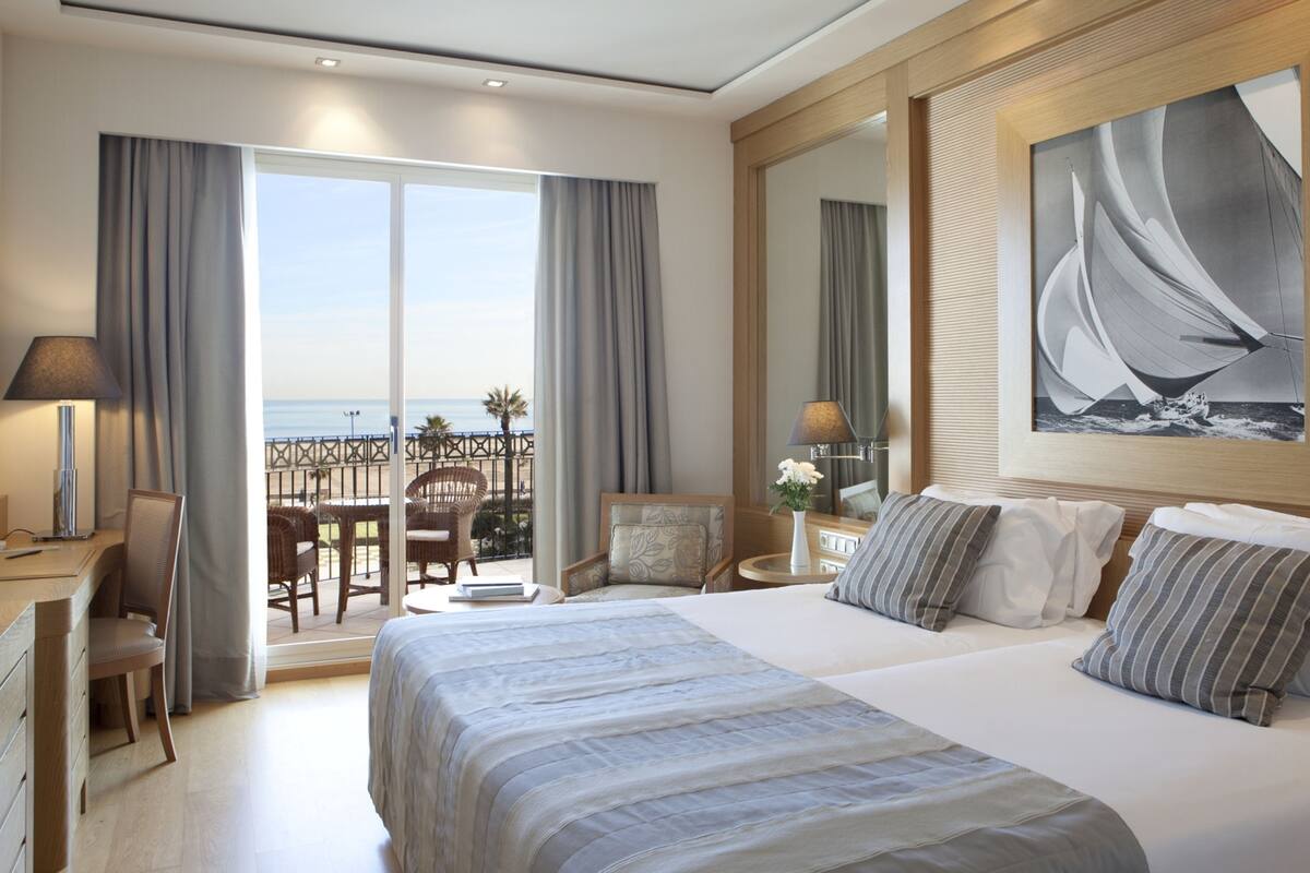 Een hotelkamer met een groot bed, een bureau, een stoel, een klein tafeltje met een lamp, en een balkon met uitzicht op de oceaan.