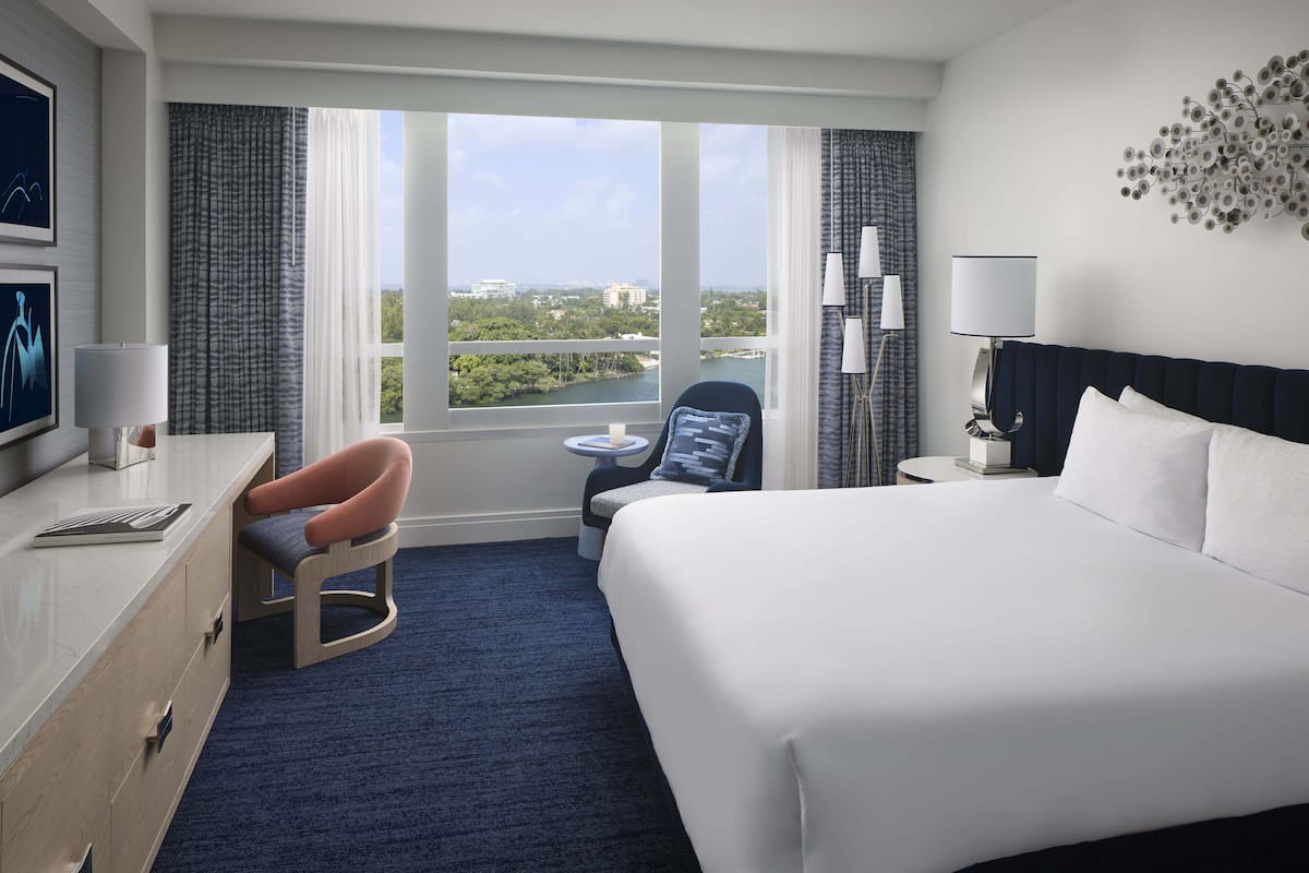 A hotel room with a large bed, a desk with a chair, a small round table, and a view of the city through large windows.