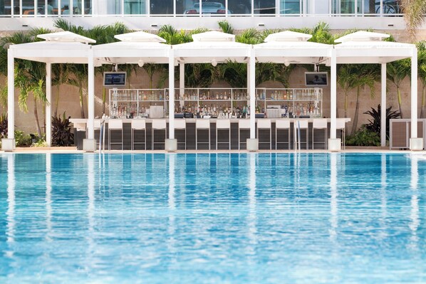 Zona de piscina con sombrillas blancas, un bar y una zona de estar con vista a un edificio al fondo.