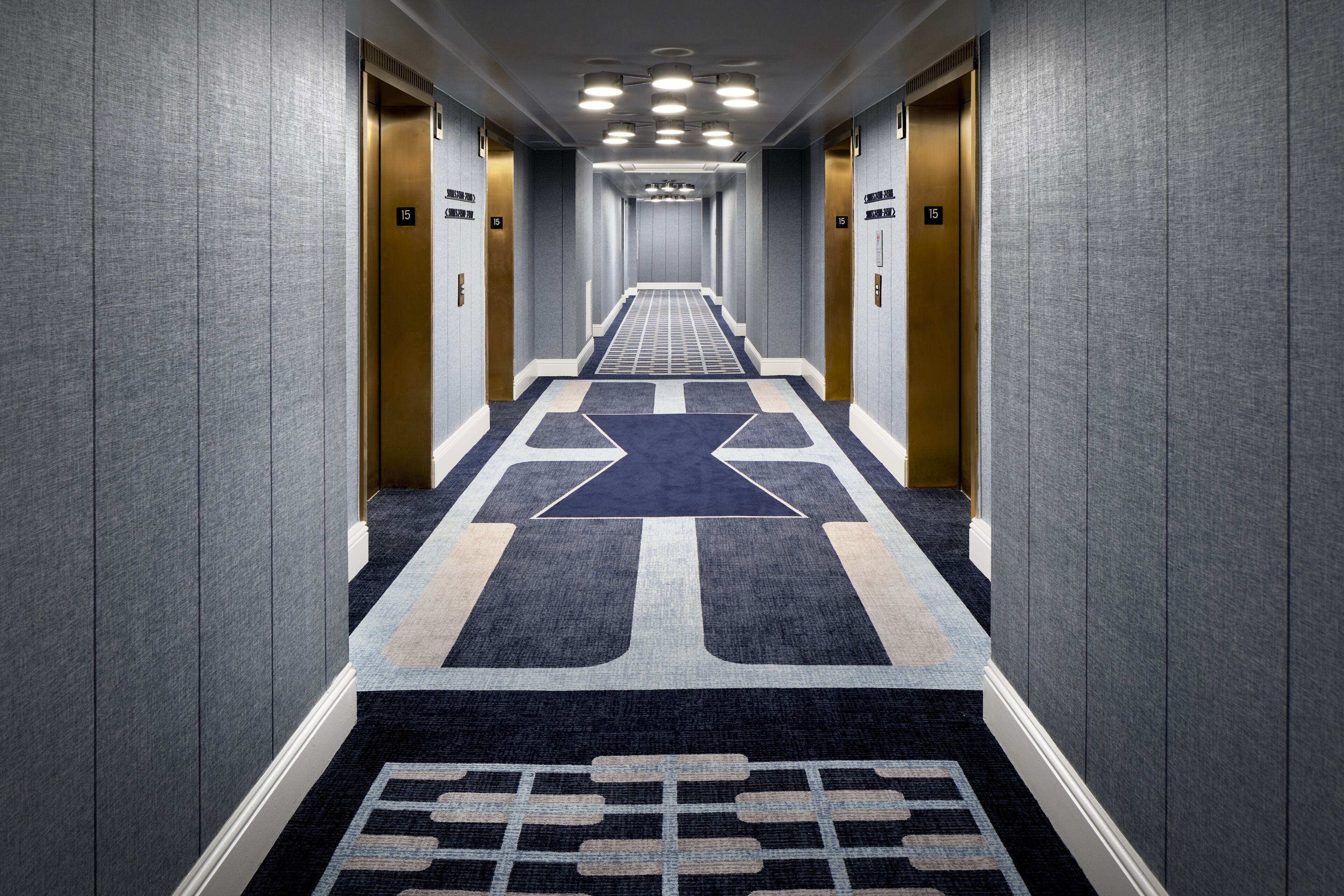 Un pasillo moderno de hotel con un diseño geométrico en la alfombra, ascensores empotrados en la pared y un color gris uniforme en las paredes.