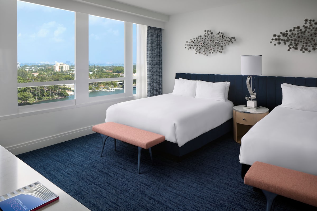 A hotel room with two beds, a bench, a nightstand, a lamp, and a view of the city.