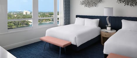 A hotel room with two beds, a bench, a nightstand, a lamp, and a view of the city.