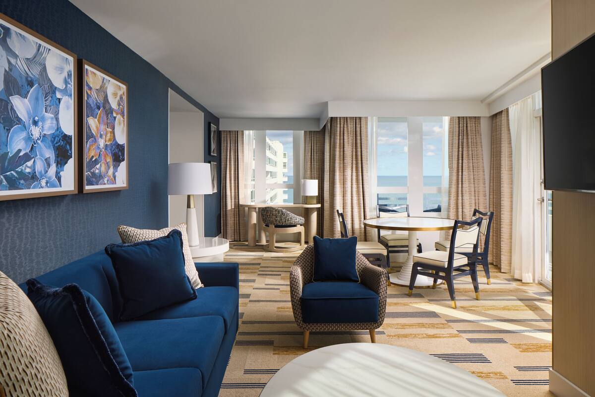 A hotel room with a blue sofa, armchairs, a dining table, and a TV.