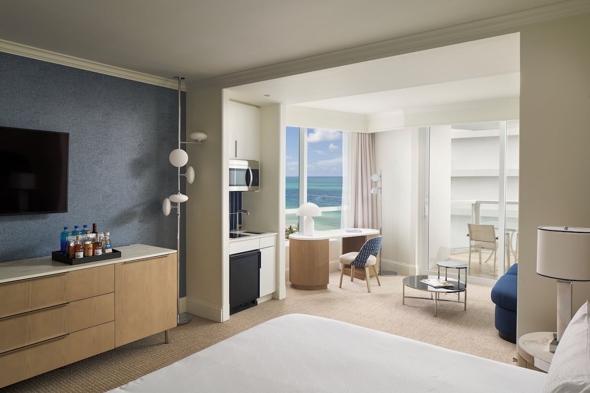 A hotel room with a bed, a desk, a chair, a small table, a TV, a minibar, and a view of the ocean.