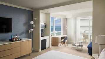 A hotel room with a bed, a desk, a chair, a small table, a TV, a minibar, and a view of the ocean.