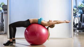 Una persona realizando un estiramiento de espalda sobre una pelota de ejercicios roja en un gimnasio con equipo de entrenamiento al fondo.