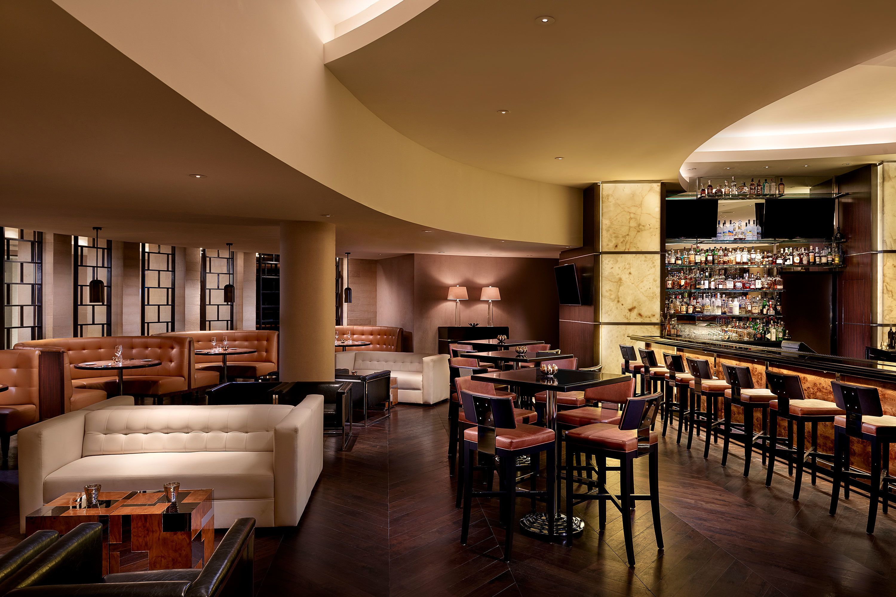 Un espace bar doté d’un long comptoir, de nombreux tabourets et d’un bar bien garni de diverses bouteilles. On y trouve également des coins salon avec des canapés et des tables, ainsi qu’un grand écran fixé au mur.