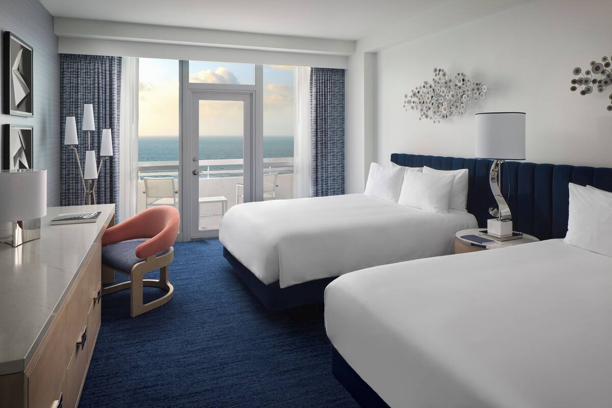 A hotel room with two beds, a desk, a chair, and a view of the ocean.