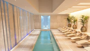 A long indoor swimming pool with a tiled floor, a metal handrail, and lounge chairs along one side.