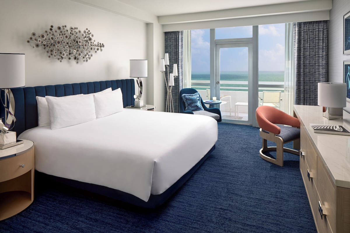 A hotel room with a large bed, a desk with a chair, a view of the ocean, and a balcony.