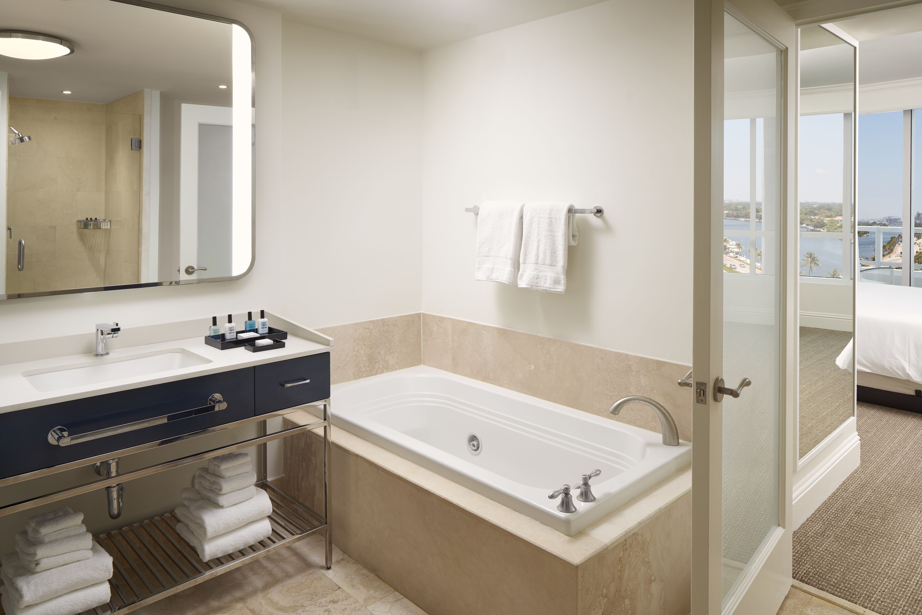 A modern bathroom with a bathtub, a large mirror, and a view of a balcony with a cityscape and water.
