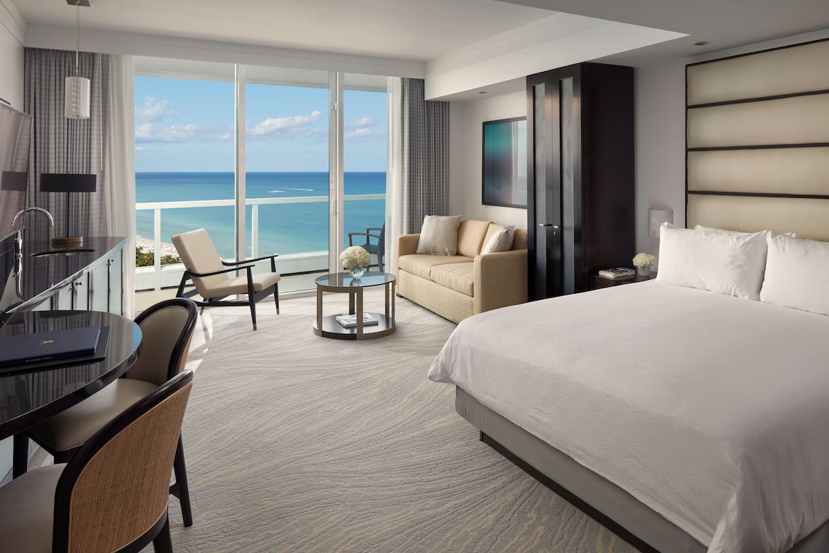 A hotel room with a large bed, a desk with a chair, a sofa, a TV, and a balcony with a view of the ocean.
