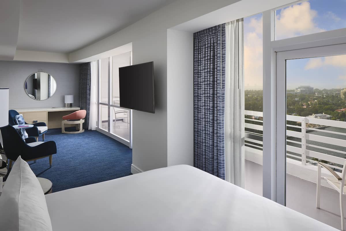 A hotel room with a bed, a desk, a chair, a TV, and a balcony with a city view.