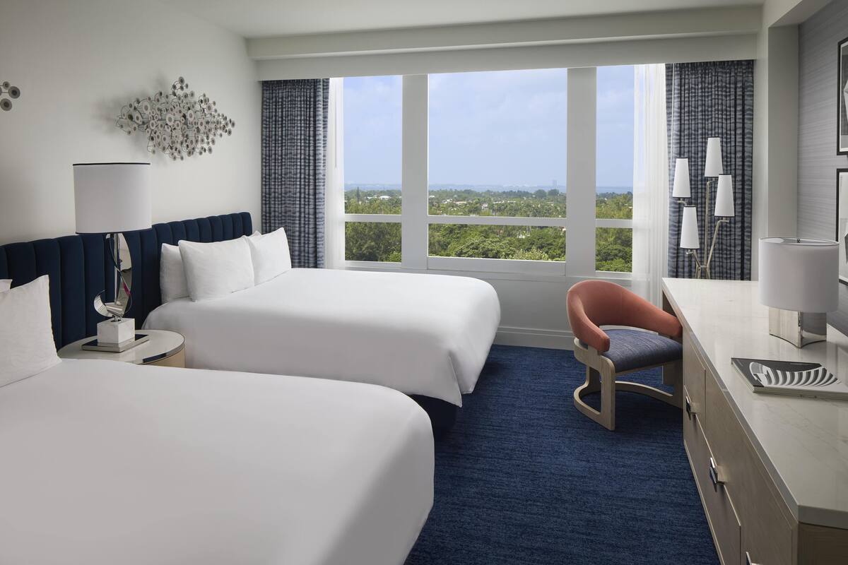 A hotel room with two beds, a desk, a chair, and a large window offering a city view.