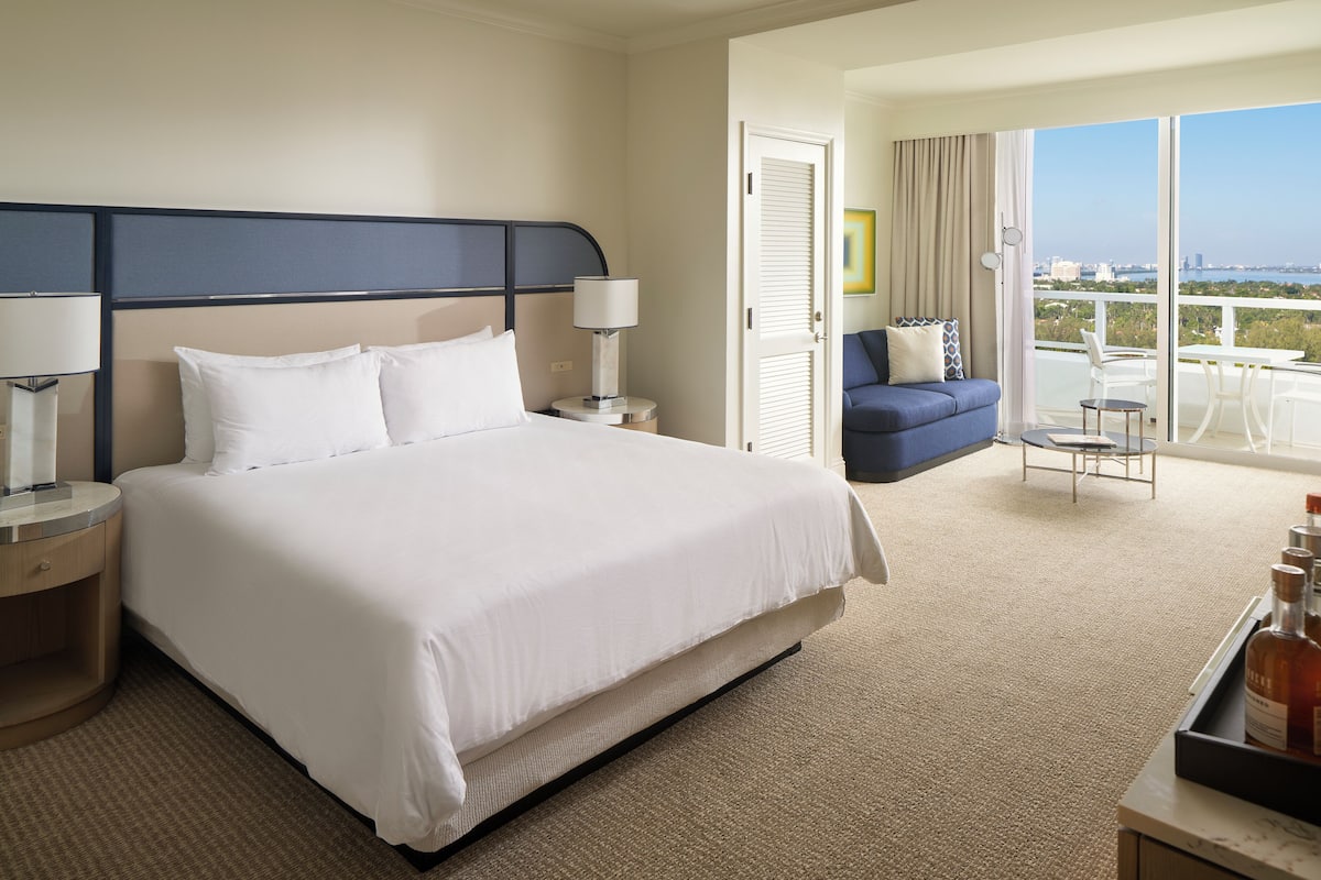 A hotel room with a large bed, a nightstand, a desk with a tray of bottles, a balcony with a view, and a blue sofa.