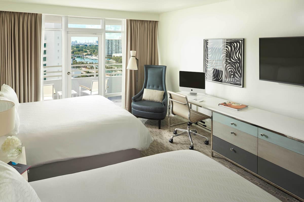 A hotel room with two beds, a desk with a computer, a chair, a TV, and a view of the cityscape.