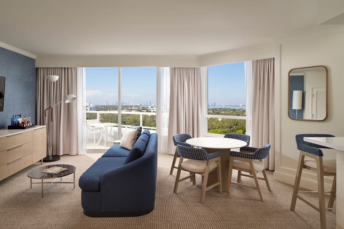 A modern hotel room with a sofa, dining table, chairs, a small coffee table, a TV, and a large window with a city view.