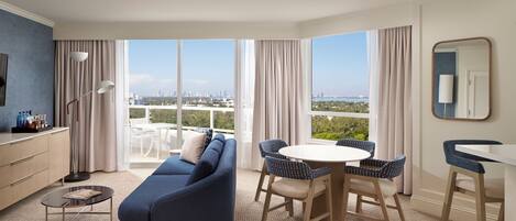 Una habitación de hotel moderna con sofá, mesa de comedor, sillas, una pequeña mesa de centro, televisor y un ventanal con vistas a la ciudad.