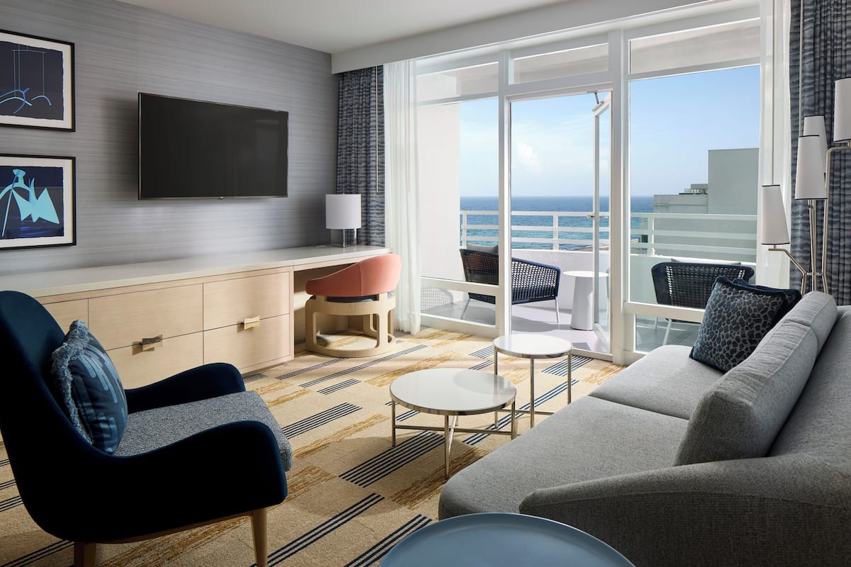 A modern hotel room with a sofa, armchair, small round table, and a TV mounted on the wall.