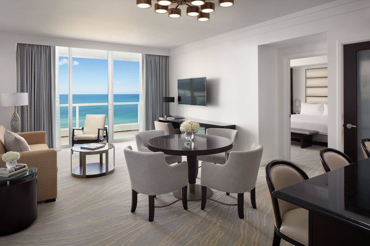 A hotel room with a round dining table, four chairs, a sofa, a TV, and a balcony with a view of the ocean.
