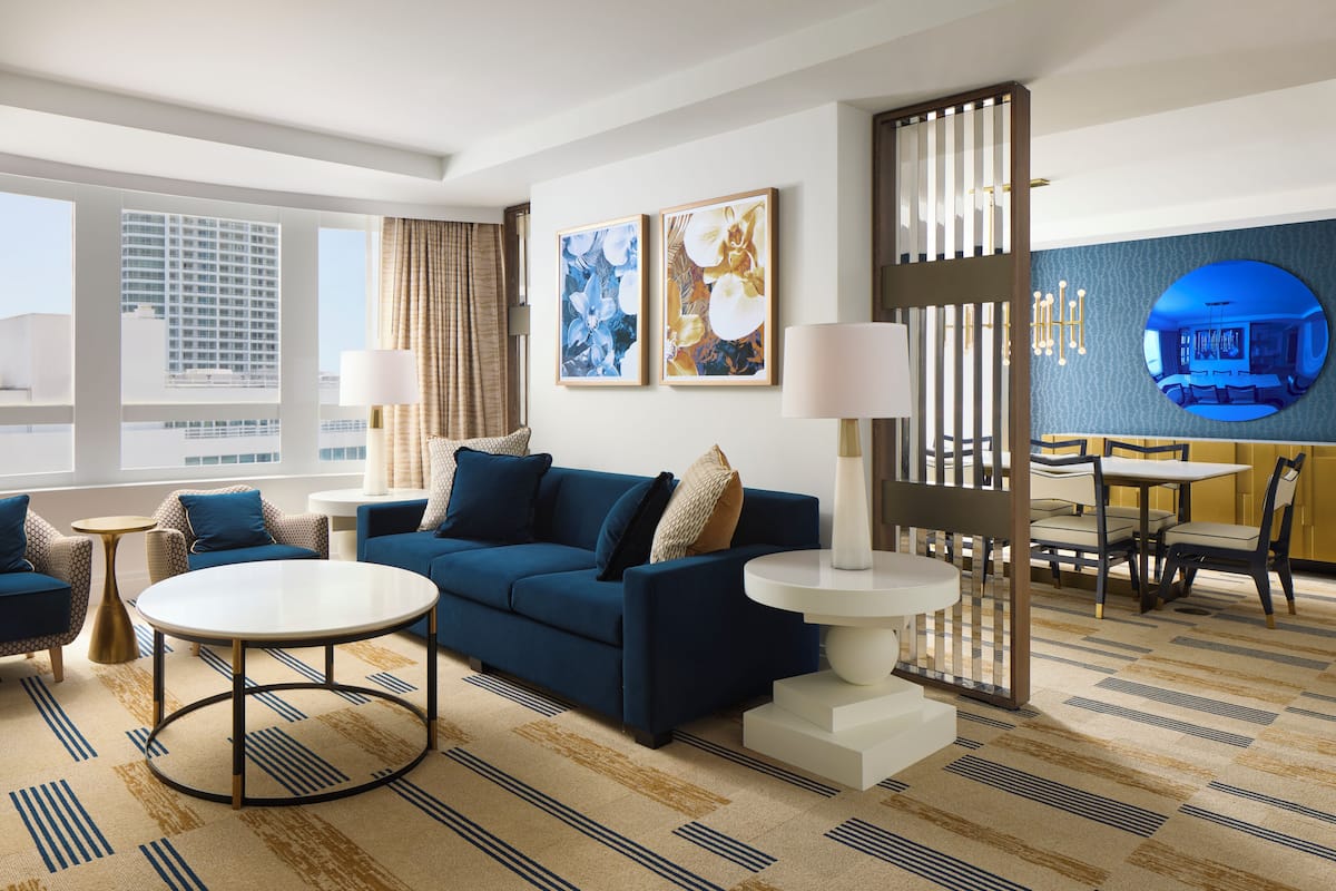 A modern hotel room with a blue sofa, a round coffee table, a dining area with a table and chairs, and large windows offering a city view.
