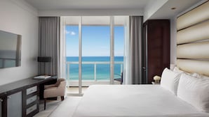 Habitación de hotel con una cama grande, un escritorio, una silla y vista al océano a través de una puerta corredera.