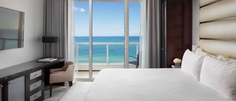 A hotel room with a large bed, a desk, a chair, and a view of the ocean through a sliding door.
