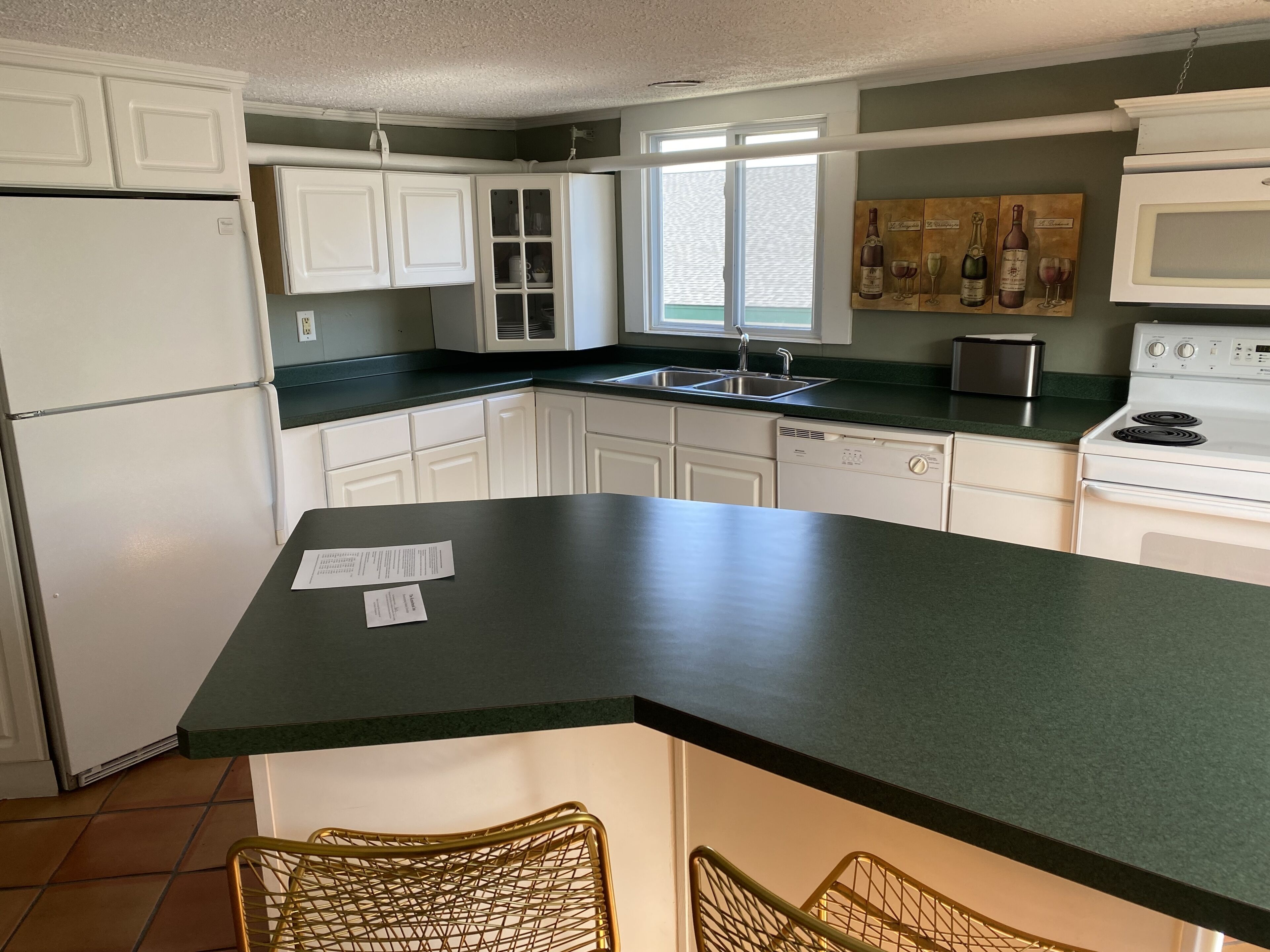 A kitchen with white cabinets, a black countertop, a window, and a painting of wine bottles.