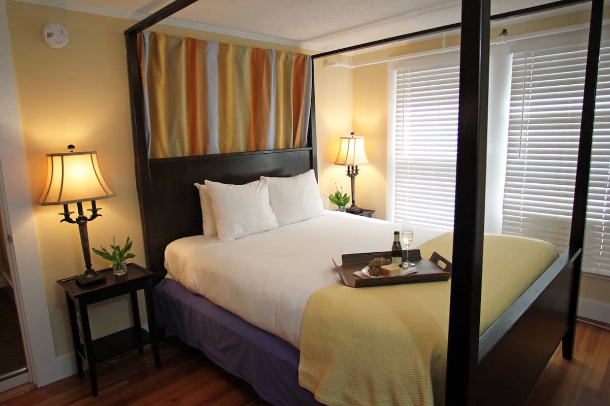 A hotel room with a large bed, two bedside tables with lamps, a window with blinds, and a painting above the bed.