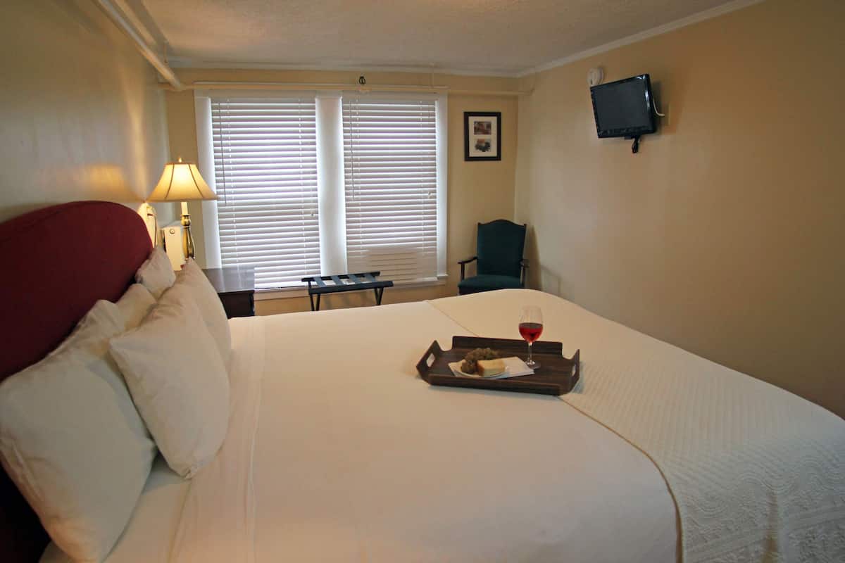 A hotel room with a bed, a bedside table with a lamp, a chair, a TV mounted on the wall, and a window with blinds.