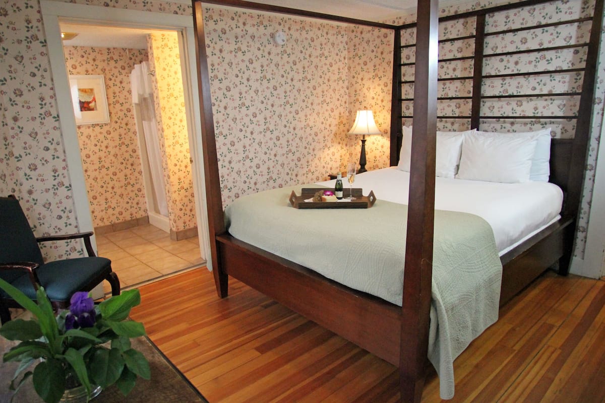 A four-poster bed with a wooden frame, a bedside table with a lamp, and a floral wallpaper pattern.