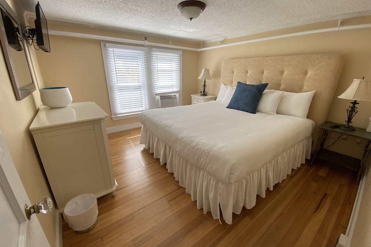 A bedroom with a large bed, a nightstand, a lamp, a window with blinds, and a ceiling fan.