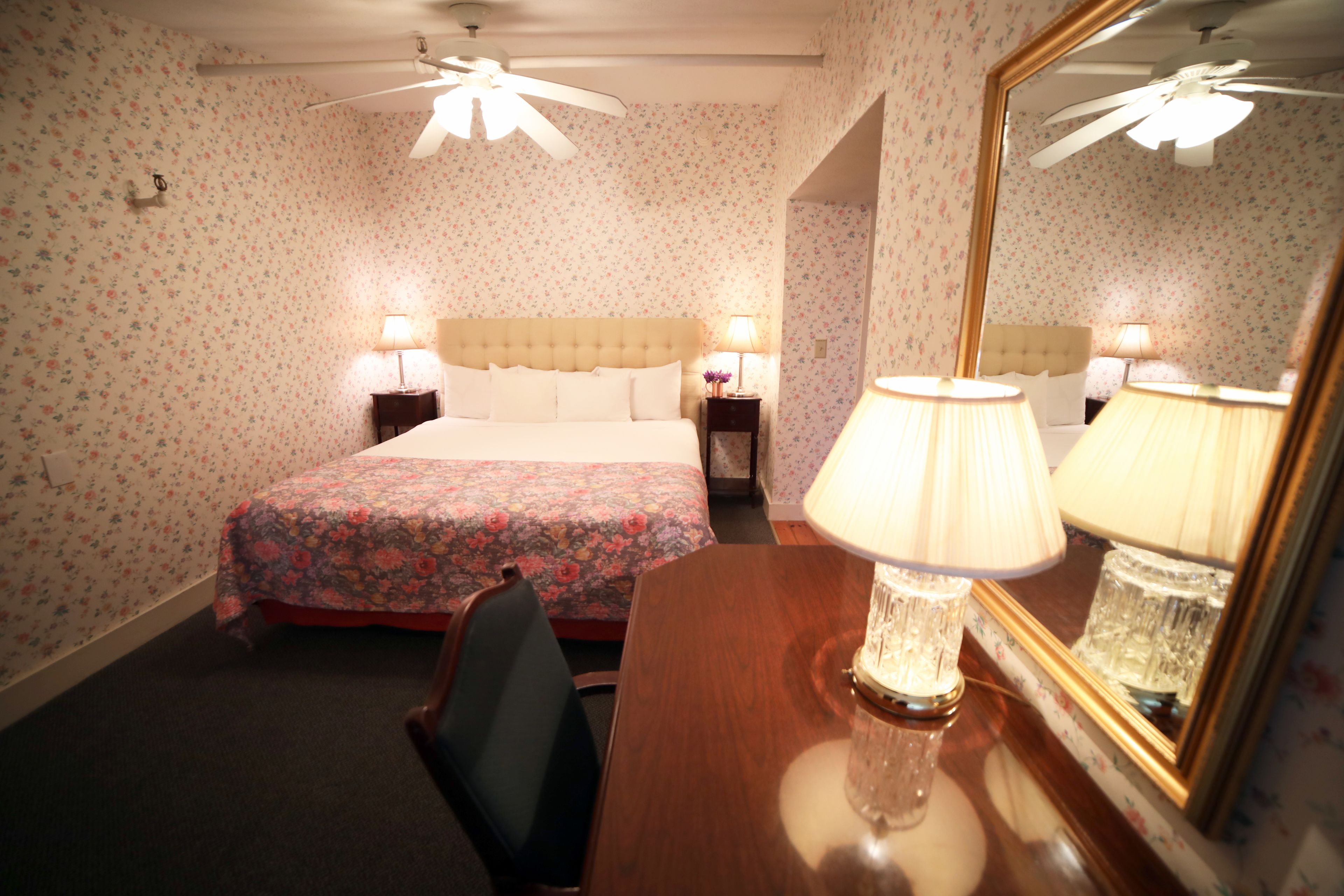 A hotel room with floral wallpaper, a bed with a floral bedspread, two bedside tables with lamps, a wooden desk with a lamp, and a ceiling fan.