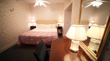 A hotel room with floral wallpaper, a bed with a floral bedspread, two bedside tables with lamps, a wooden desk with a lamp, and a ceiling fan.