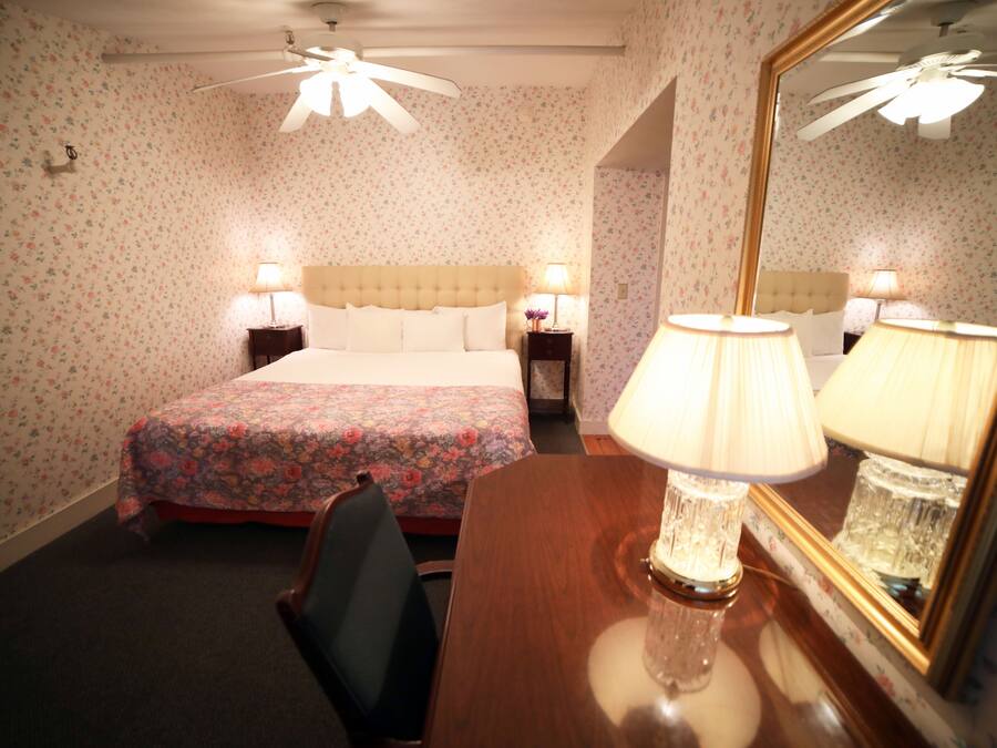 A hotel room with floral wallpaper, a bed with a floral bedspread, two bedside tables with lamps, a wooden desk with a lamp, and a ceiling fan.