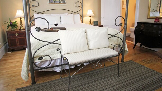 Una cama con dosel, con cubrecama y almohadones blancos, flanqueada por dos mesitas de noche con lámparas, un aparador y un espejo en la pared.