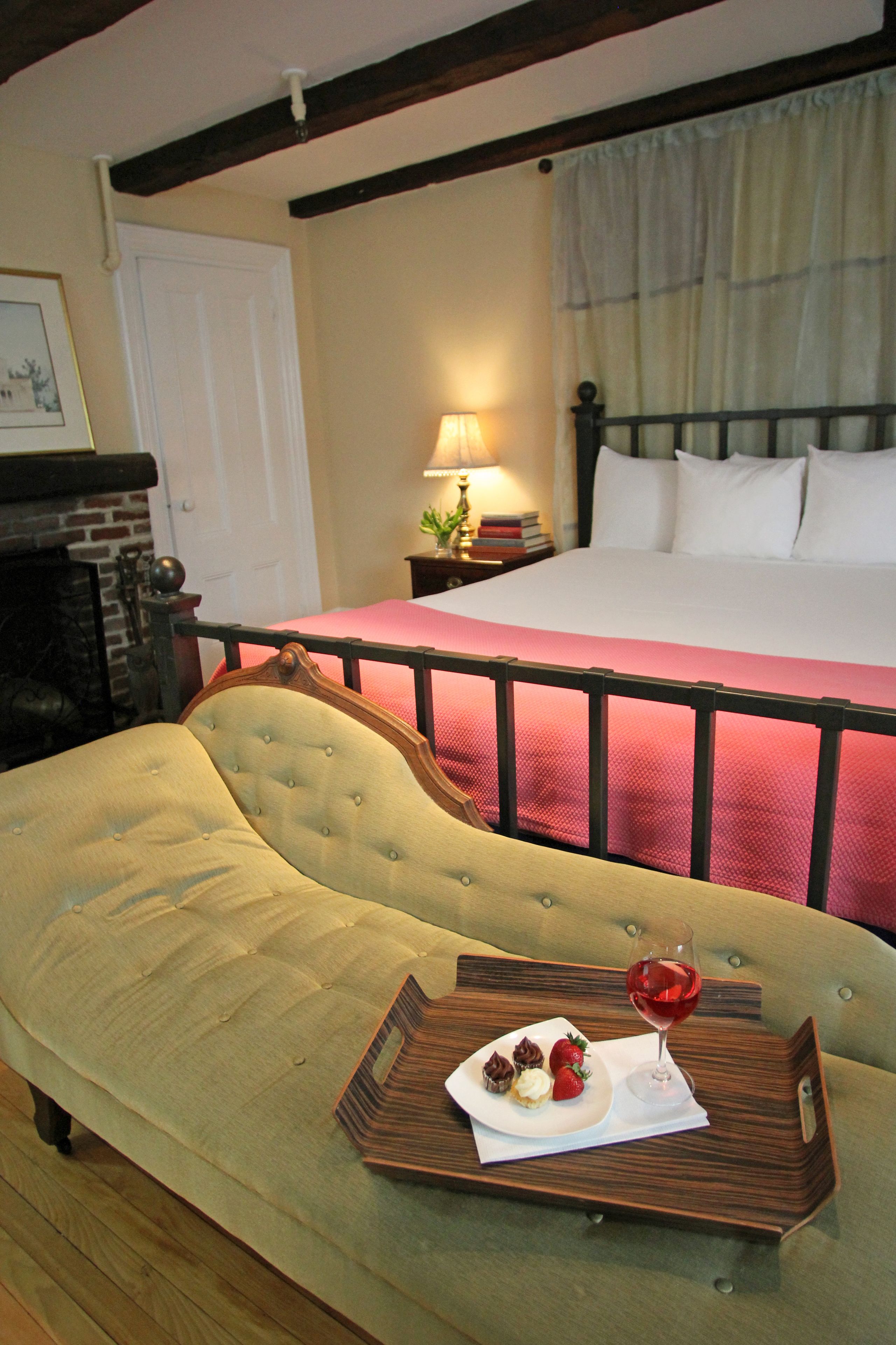 A hotel room with a bed, a sofa, a tray with desserts and a glass of red wine, and a fireplace.