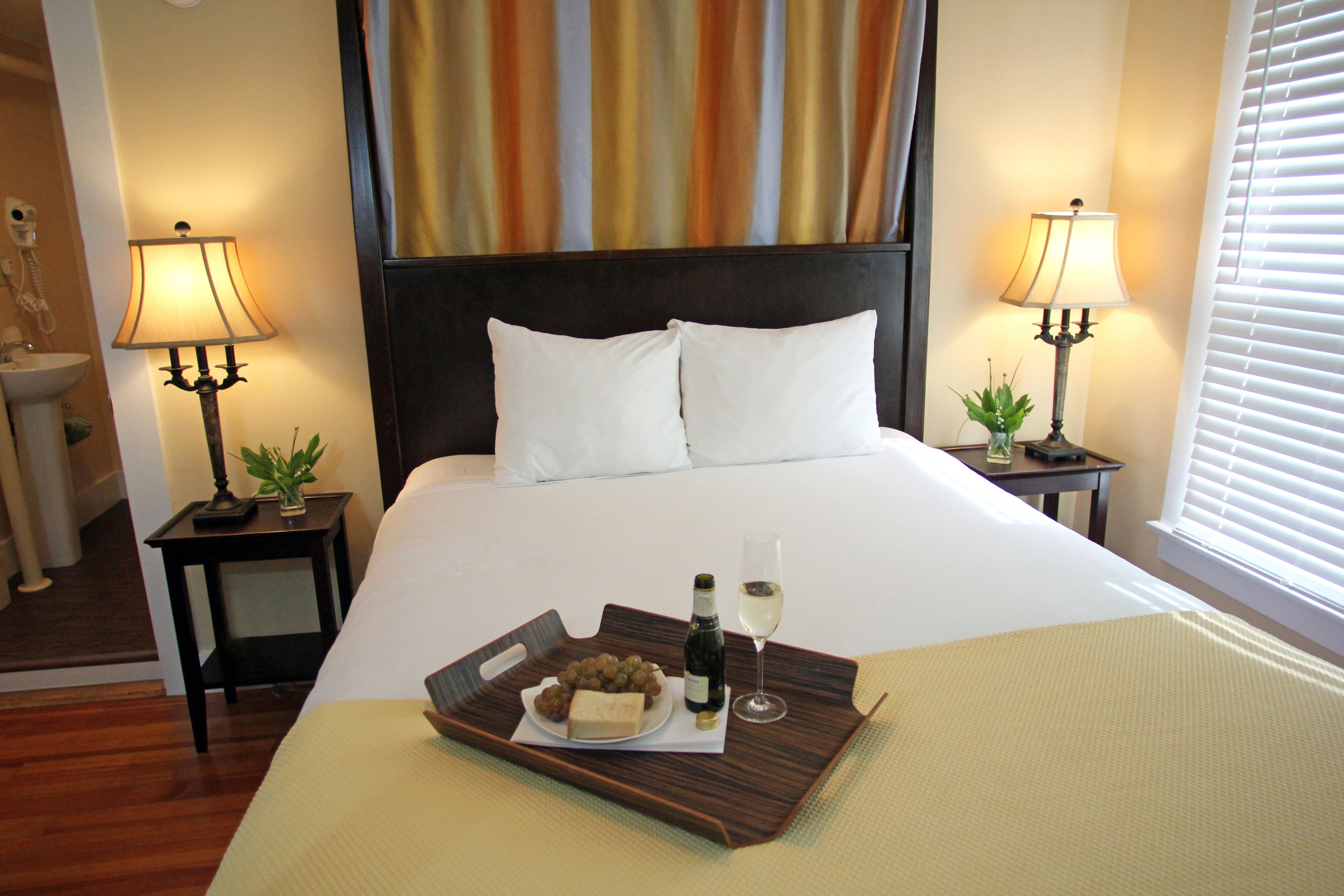A hotel room with a large bed, two bedside lamps, a tray with food and drinks, and a window with blinds.