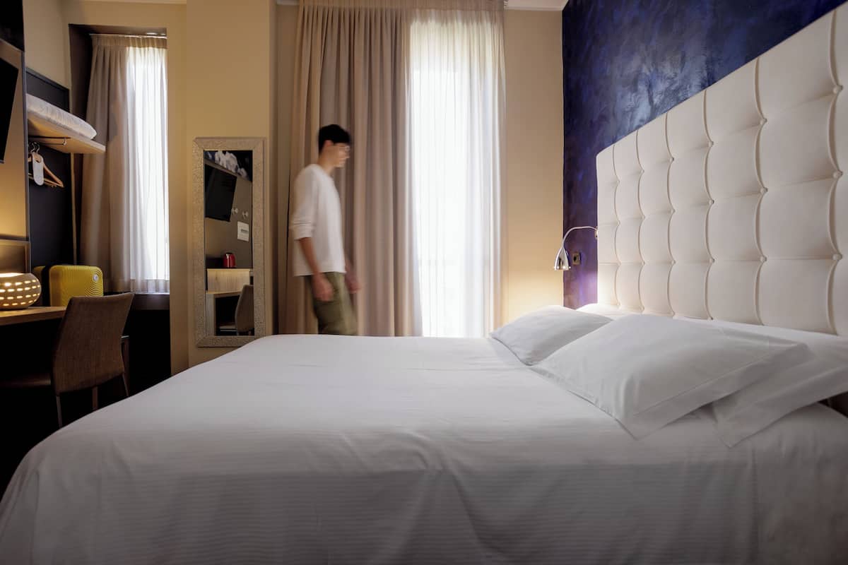 A hotel room with a large bed, a desk, a chair, a mirror, and a window with curtains.